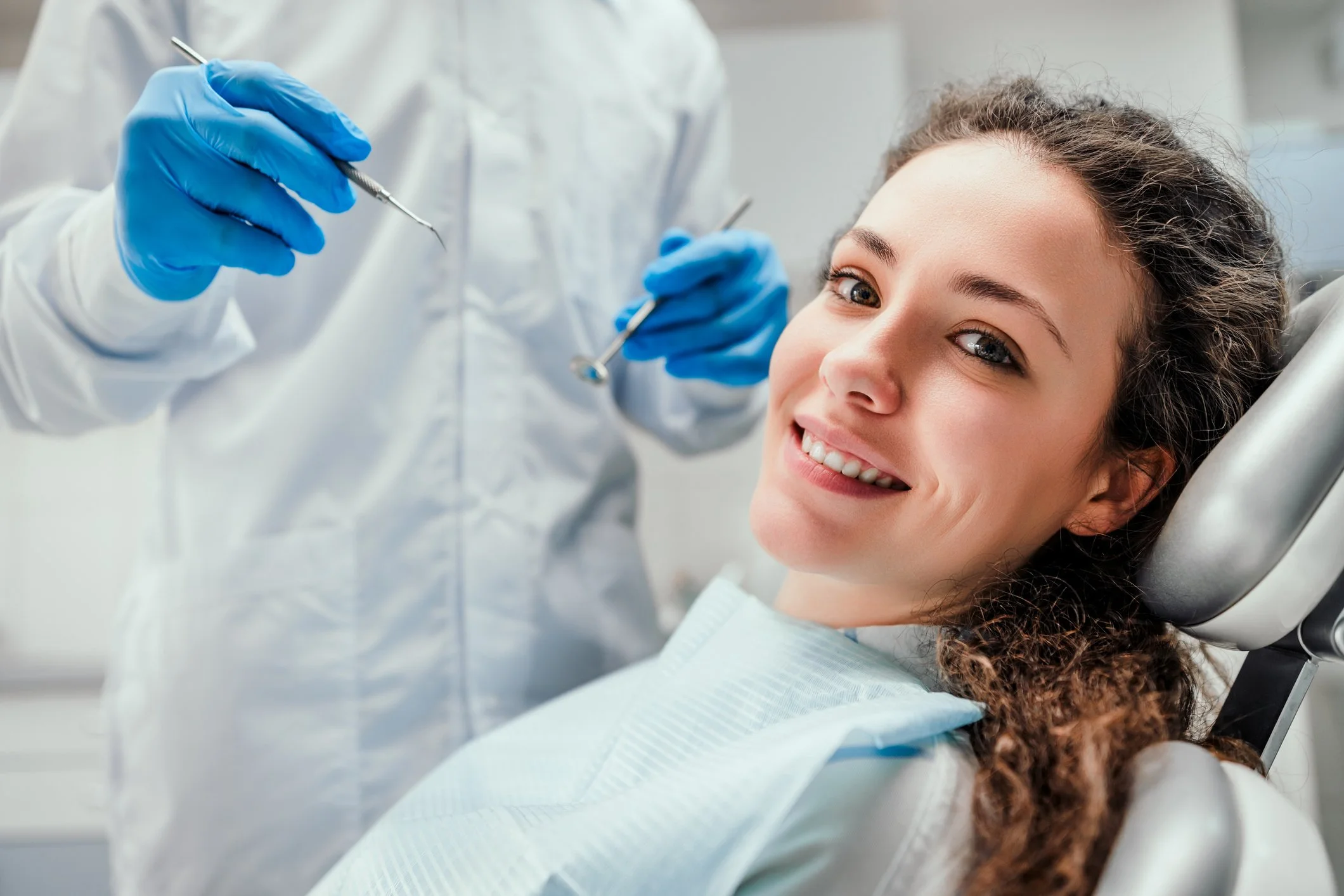 A young woman with curly brown hair smiling while lying in a dental chair at the dentist's office, wearing a light blue dental bib.