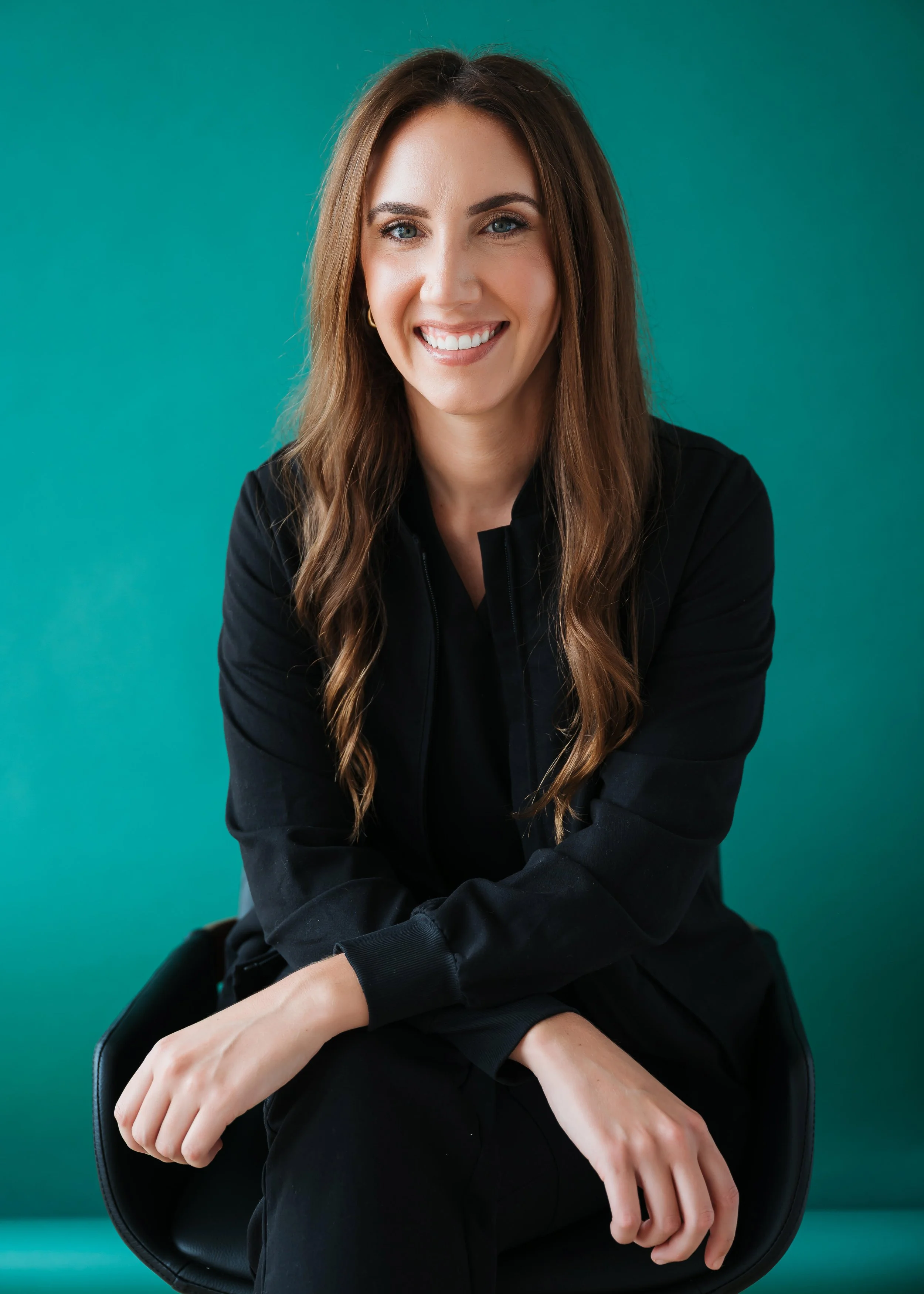 A young woman with long brown hair and blue eyes smiling, sitting in a black chair against a teal background, wearing a black top.