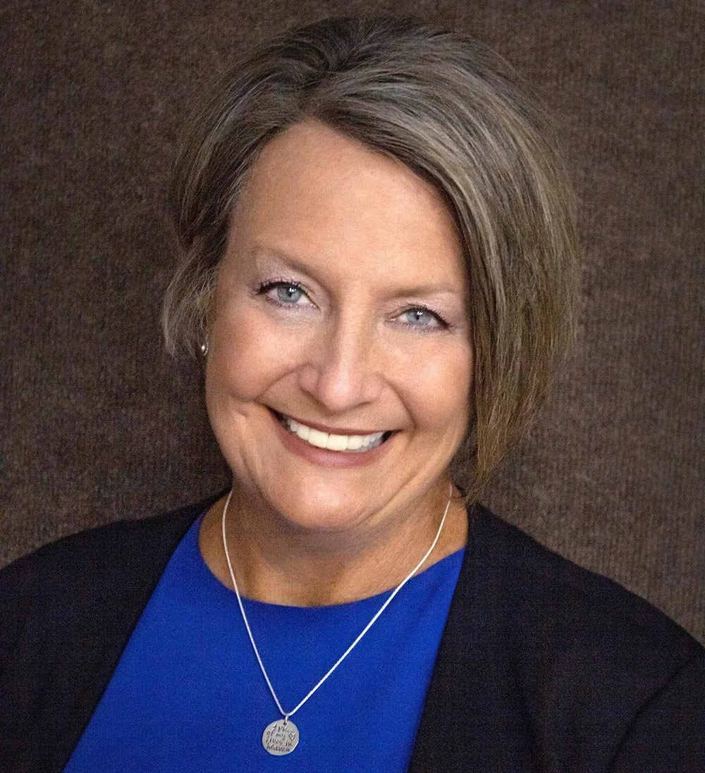 A smiling woman with short, light brown hair and blue eyes wearing a blue top, black cardigan, and a round necklace, against a brown background.
