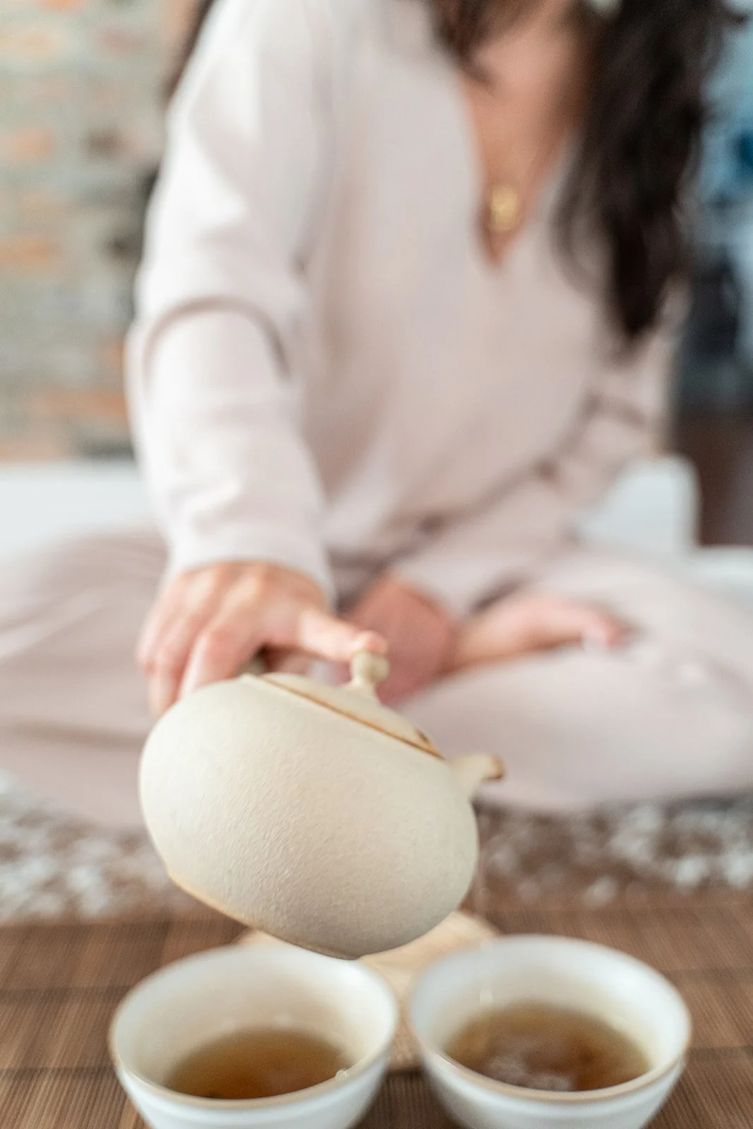 close up of a ceramic beige tea pot being poured into two cups by a woman sitting cross legged shoulders down by photographer Lenee Love