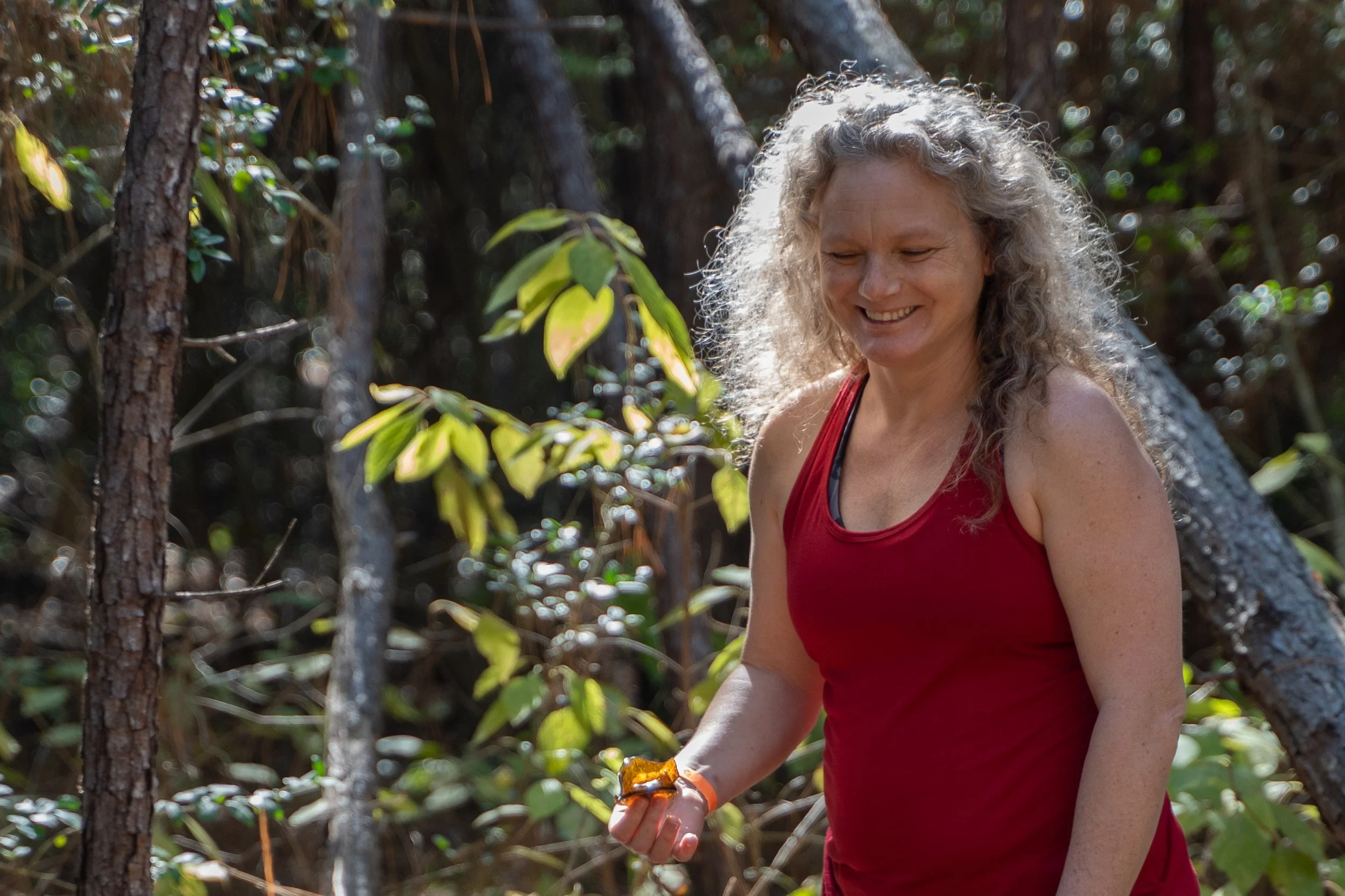 woman with curly grey hair and a red tank top holds a colorful leaf in the woods smiling by photographer Lenee Love