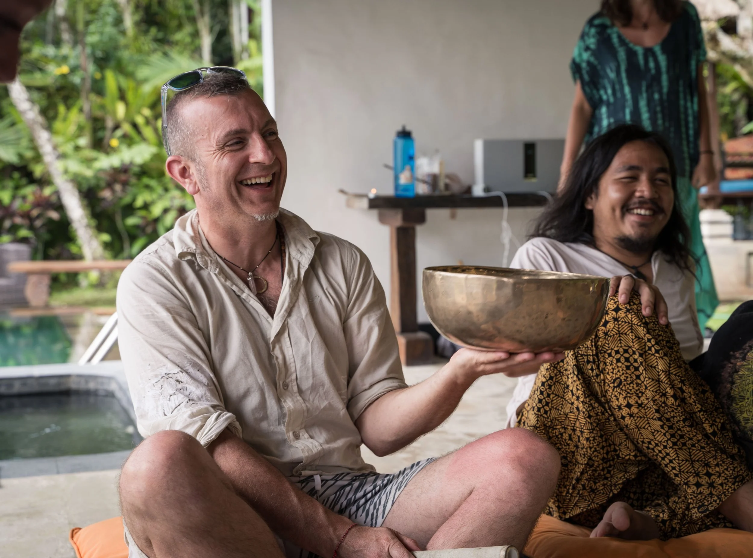 man smiling holding a tibetan singing bowl by the pool at a retreat in Bali by photographer Lenee Love