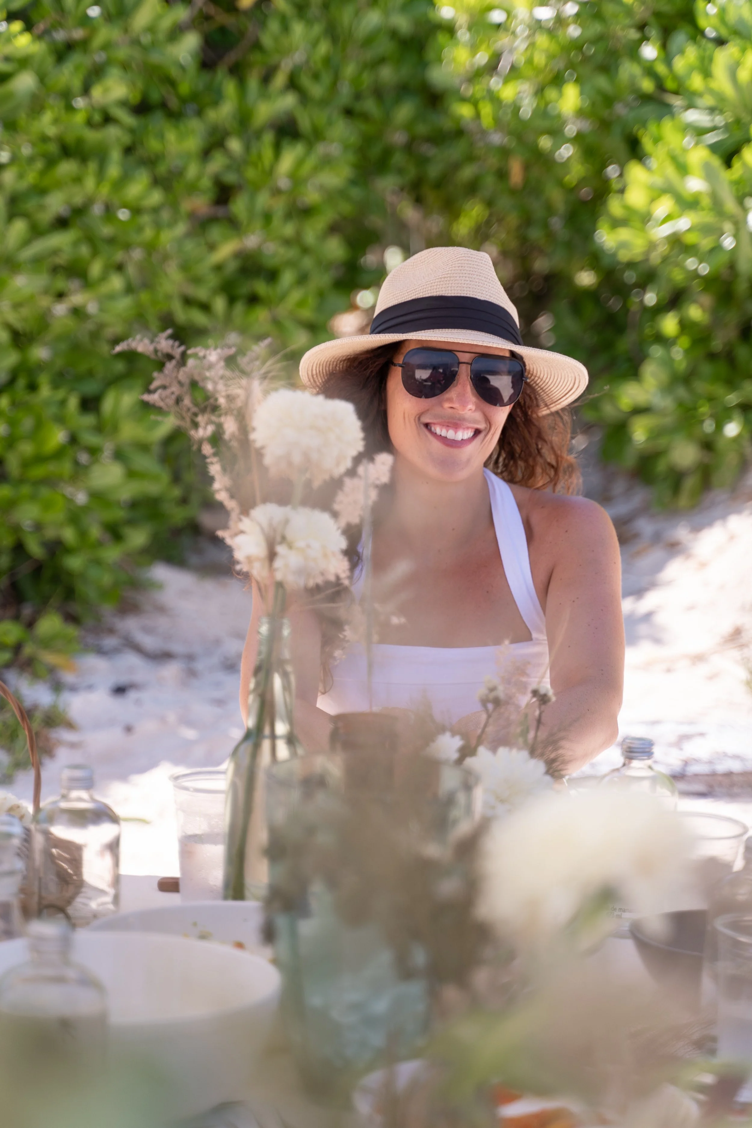 woman in white top straw hat and sunglasses smiles at the head of a picnic table on the beach with flowers in the foreground and shrubs in the background by retreat photographer Lenee Love
