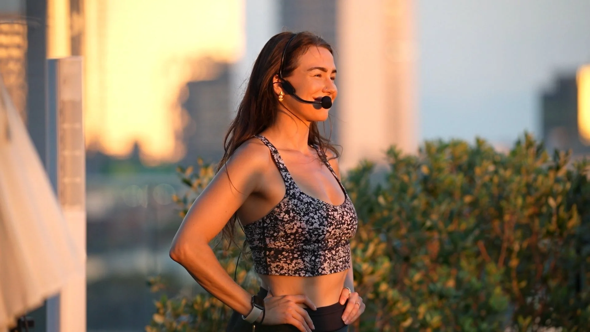 A female yoga teacher wearing a lululemon set, standing outdoors during sunset, with a headset microphone, smiling and looking towards the distance.