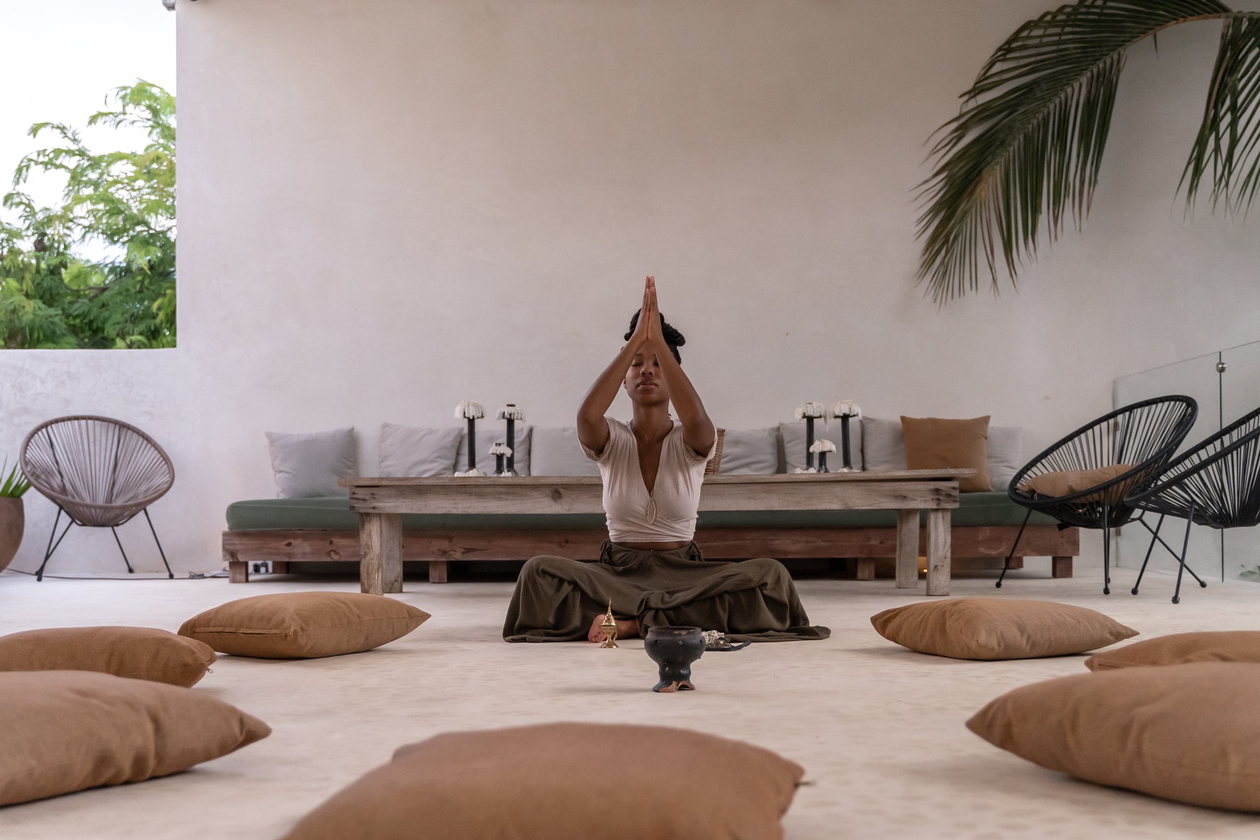 woman in green linen pants and a beige top seated on a brown cushion with prayer hands at her forehead in tulum by by retreat photographer Lenee Love
