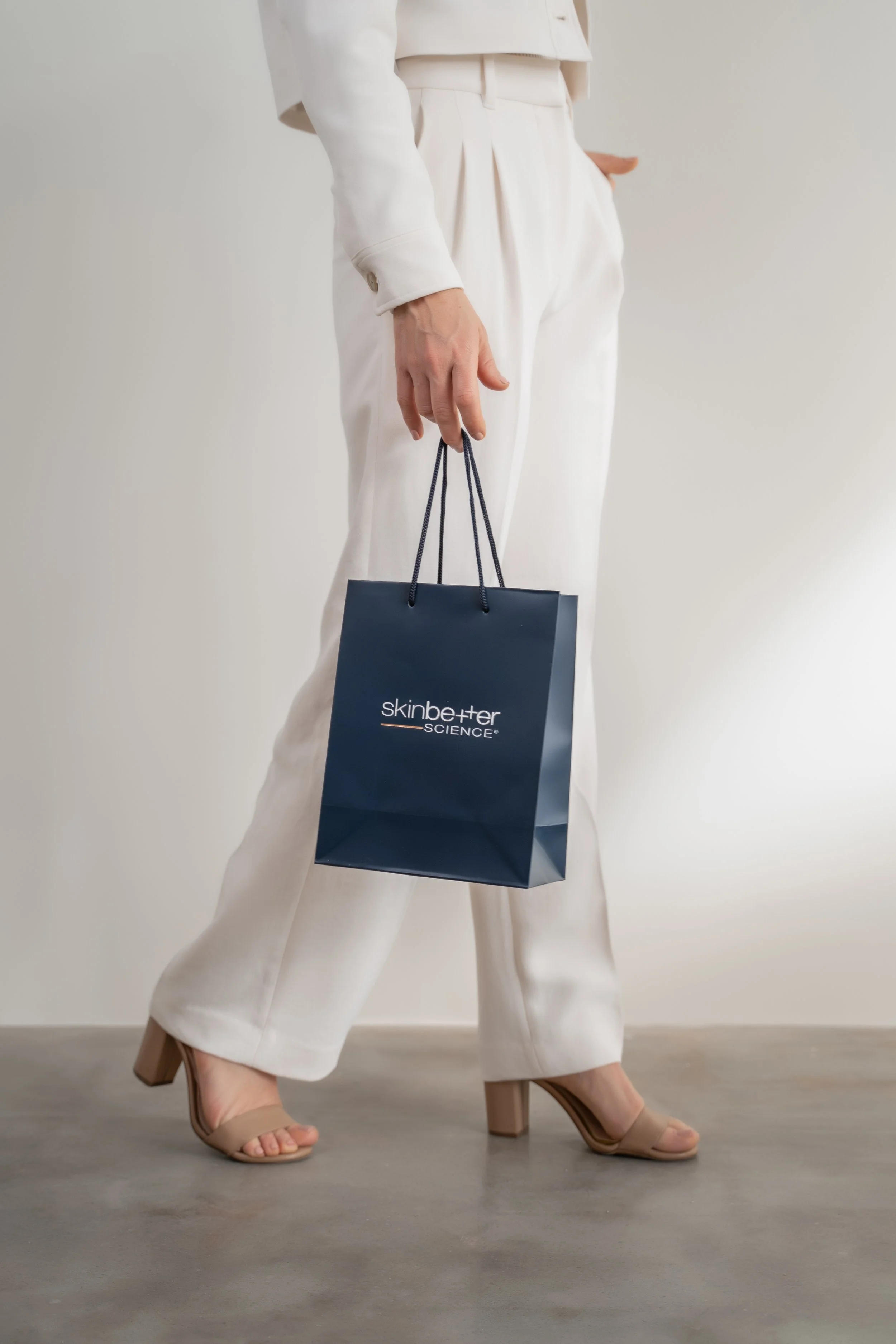 woman in white dress pants and cropped jacket beige heels walking with a skin better science bag editorial by product photographer Lenee Love