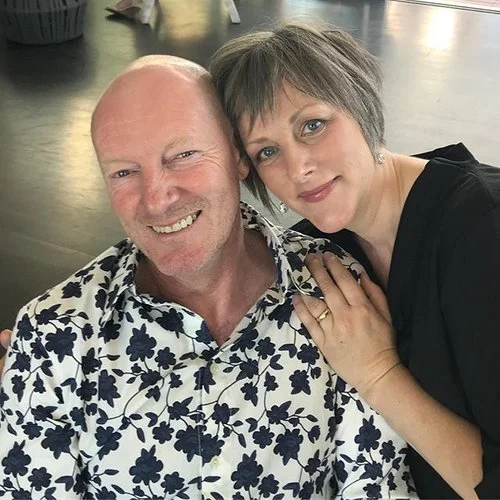 A smiling couple posing together, the man wearing a floral patterned shirt and the woman leaning on his shoulder, indoors.