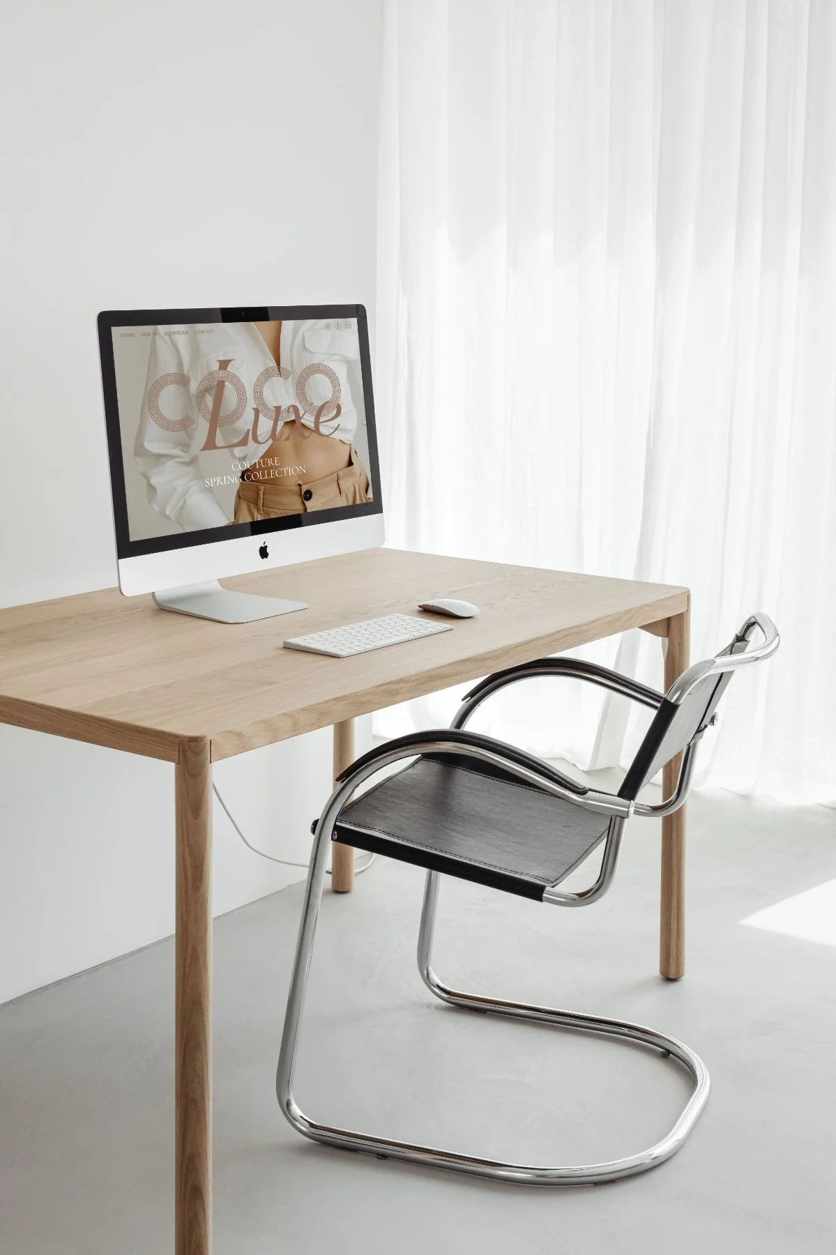 Minimalist home office setup with a wooden desk, computer monitor displaying "Coco" website, keyboard, mouse, and a modern black and chrome chair, against a white curtain background.