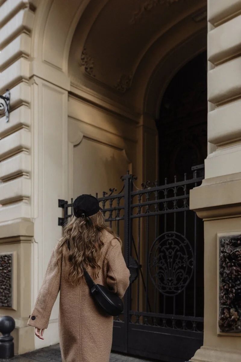 Woman in wool coat and black hat approaching ornate iron gate entrance