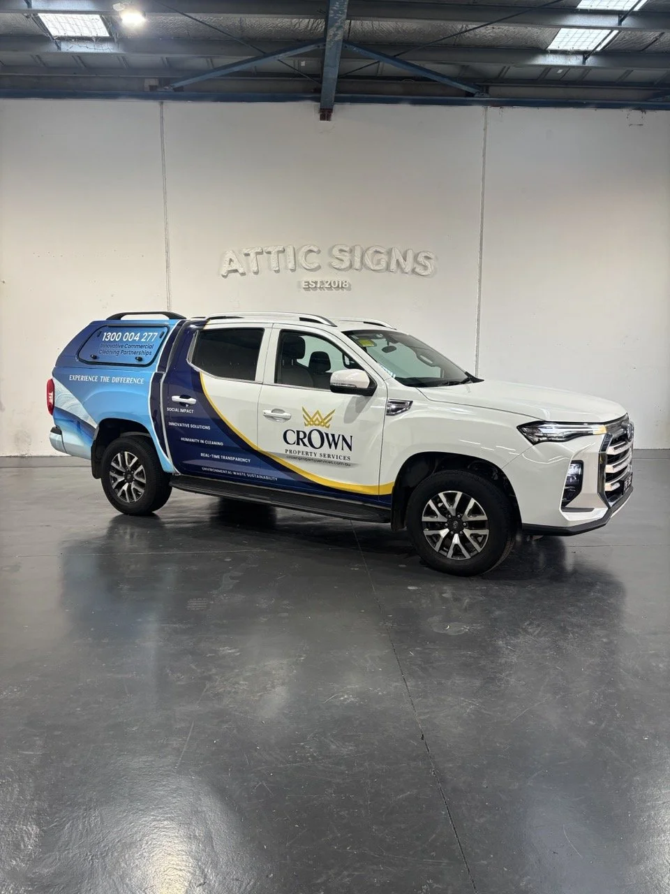 White pickup truck with blue, black, and yellow branding for Crown Property Services parked inside a warehouse with a concrete floor and a sign on the wall that reads "ATTIC SIGNS latest2018."