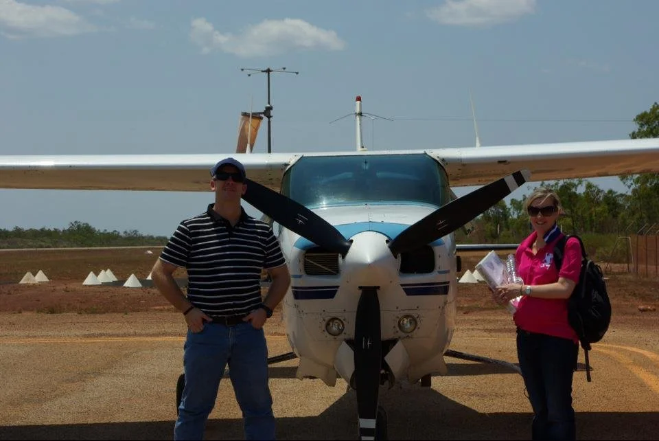 A man and a woman standing in front of a small white and blue airplane at an outdoor airport or airstrip, with some trees and a partly cloudy sky in the background.