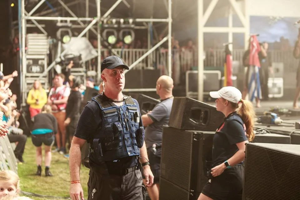 A staff member in black tactical vest and cap watches at a concert or festival. People are seen in the background, some sitting and some standing near the stage area with lighting and sound equipment.