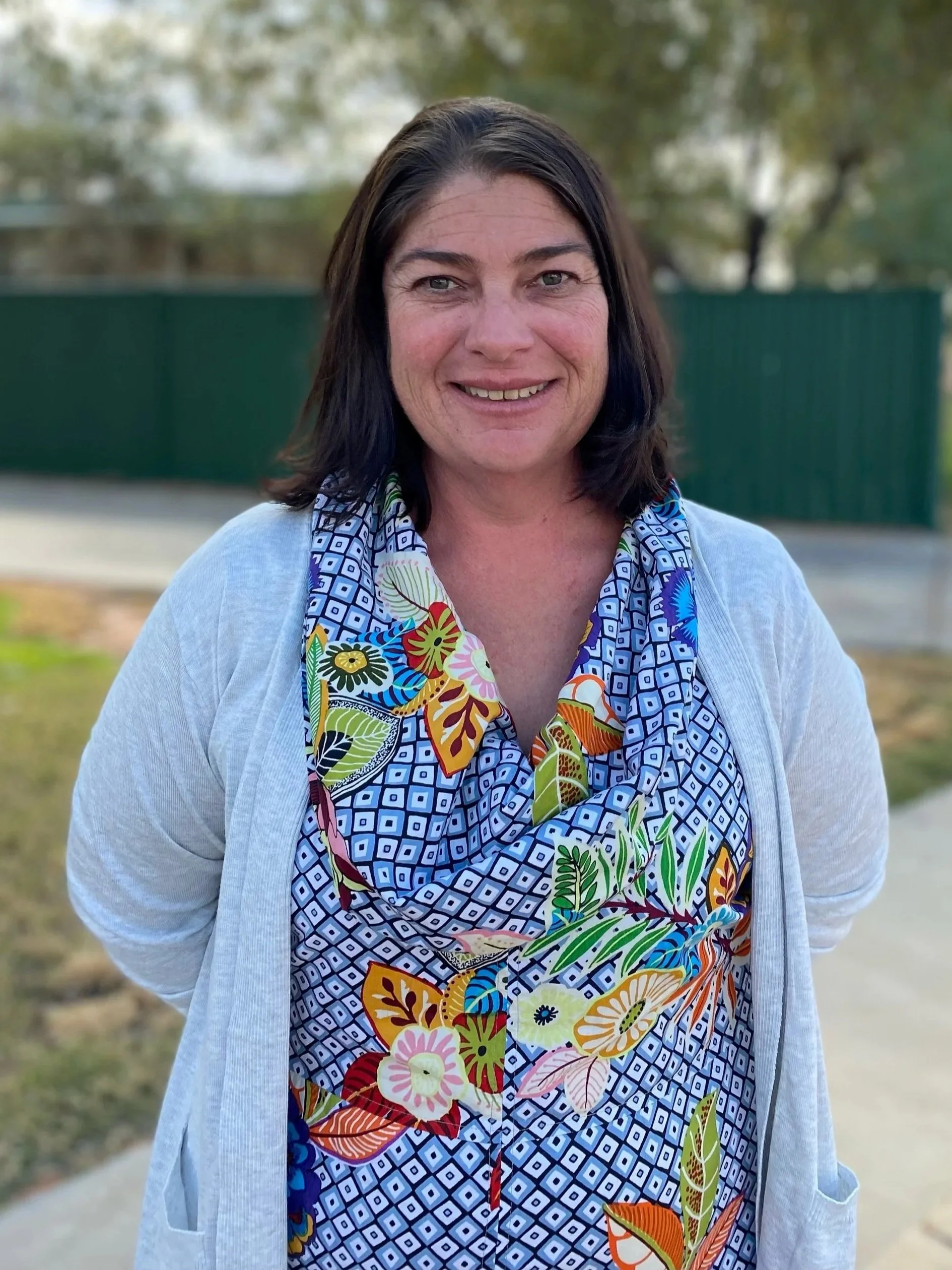 A woman with dark hair, smiling, wearing a colorful floral-patterned scarf and a light gray cardigan, standing outdoors with blurred trees and a green fence in the background.