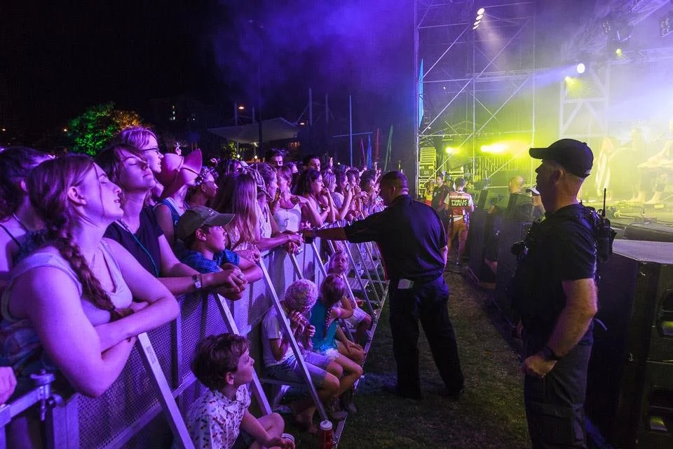 Crowd of children and adults watching a concert or event at night, with security personnel and stage lights.