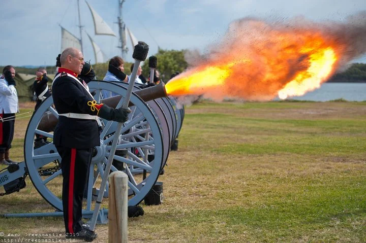 A historical reenactment with individuals dressed in 19th-century military uniforms firing a cannon on a grassy field near a river, with a large plume of fire and smoke emanating from the cannon's mouth.