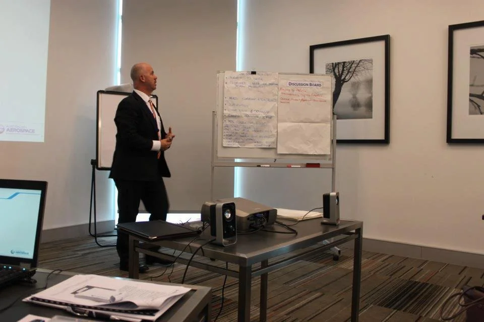 A man in a suit presenting in a conference room with a whiteboard, projector, and framed black-and-white landscape photos on the wall.