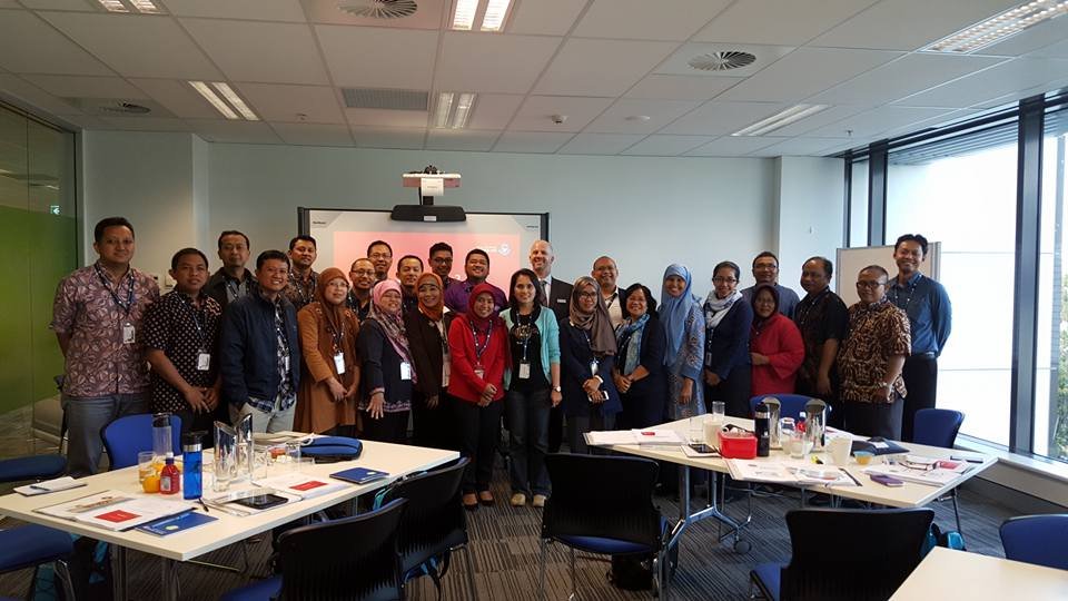 Group of diverse professionals in a meeting room posing for a photo after a workshop or seminar, with tables and materials in the foreground and a presentation screen in the background.