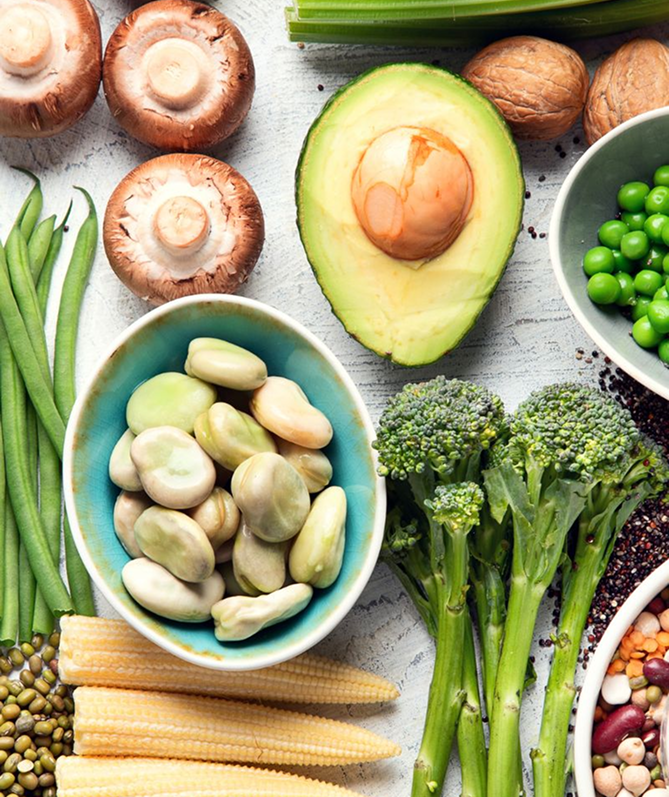 A variety of fresh vegetables including mushrooms, avocado, green beans, kidney beans, broccoli, baby lima beans, baby corn, green peas, and walnuts arranged on a light surface.