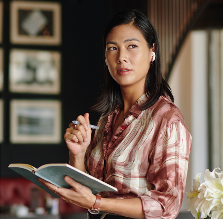 A woman holding a pen and an open notebook, sitting indoors near large windows, with framed pictures on a dark wall behind her.