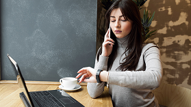 Woman sitting at a table with a laptop, coffee cup, and smartphone, looking at her phone contemplatively in a cafe or workspace.