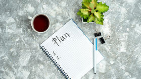 A planner, a pen, a cup of coffee, and a small plant on a gray textured surface.
