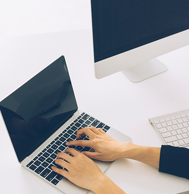 Person using a laptop on a white desk with a large monitor and keyboard nearby.