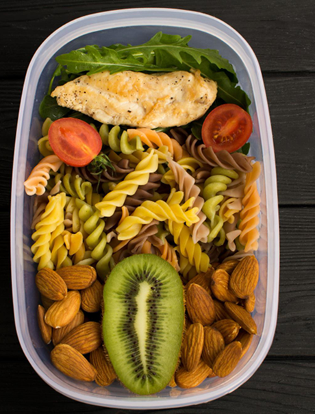 A bento box with grilled chicken, cherry tomatoes, leafy greens, multicolored rotini pasta, a halved kiwi, and almonds.