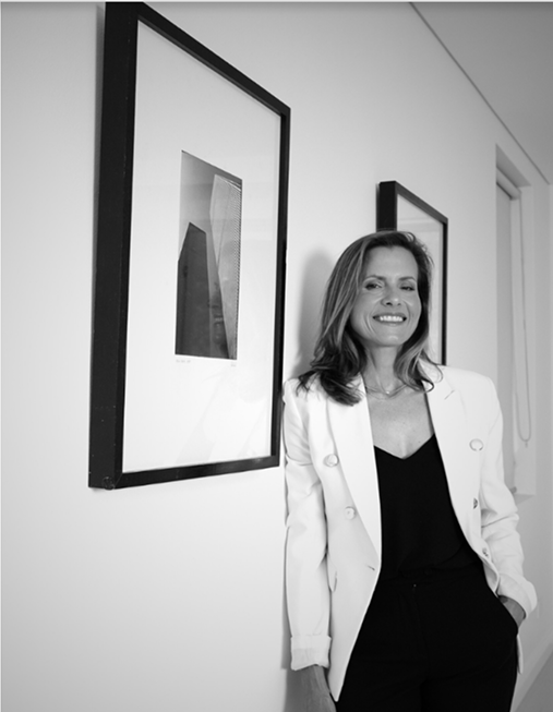 A woman with long hair smiling, dressed in a black top and white blazer, standing next to framed artwork on a white wall.