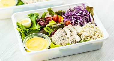 Salad in a white container with lettuce, cherry tomatoes, purple cabbage, grilled chicken, couscous, and a small cup of dressing.