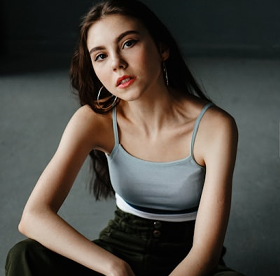 Young woman with long brown hair wearing a light blue tank top and black pants, sitting against a dark background.