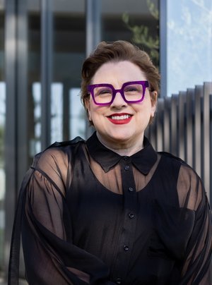 Smiling woman with short hair, wearing large purple glasses and a sheer black blouse, standing outdoors in front of a modern building.