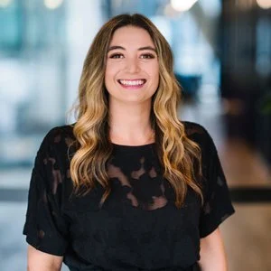 A woman with long wavy hair and a big smile, wearing a black lace top, standing indoors with a blurred background.