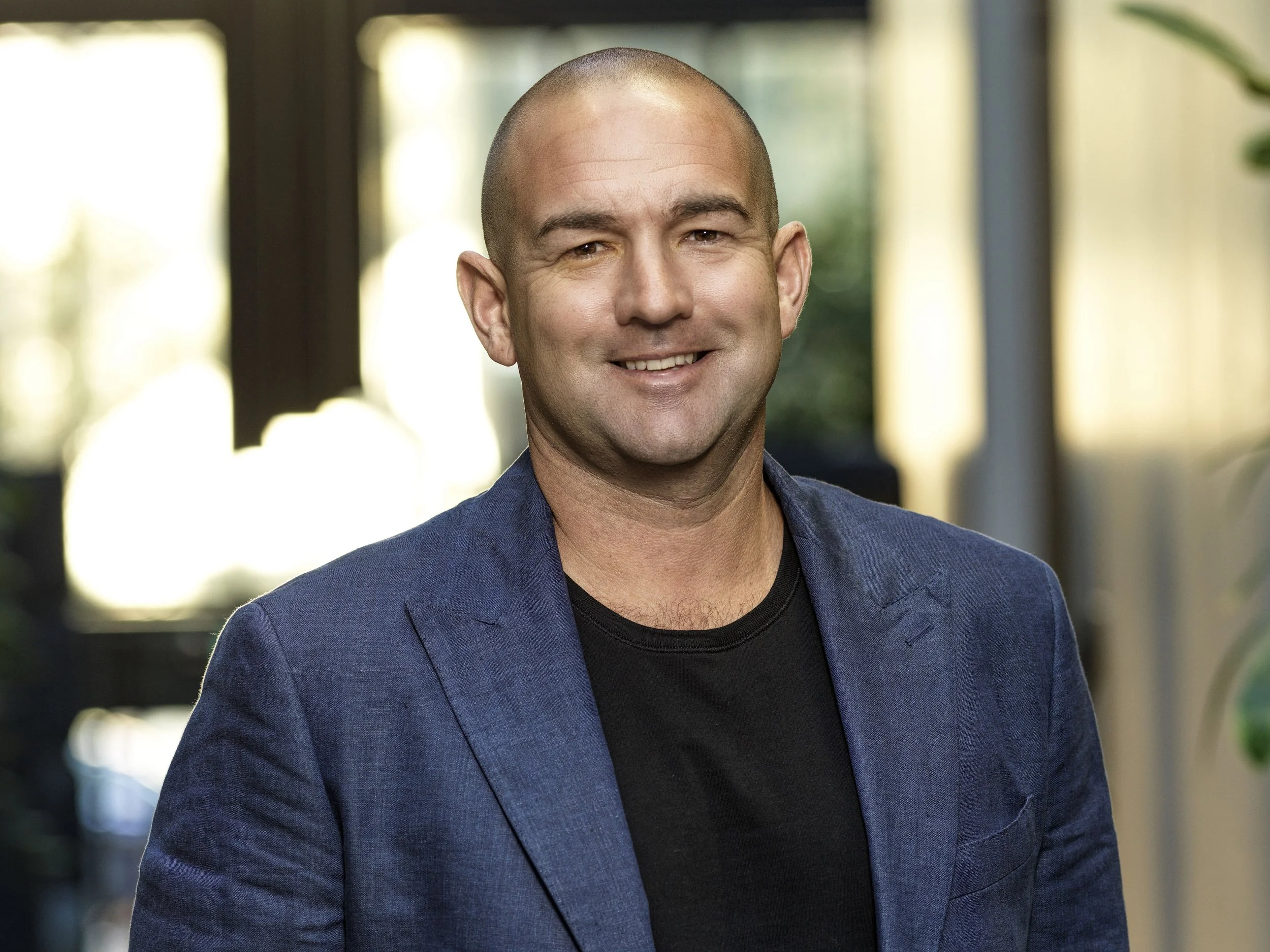 A man with a shaved head smiling, wearing a blue blazer and black shirt, standing indoors with blurred background lighting and greenery.