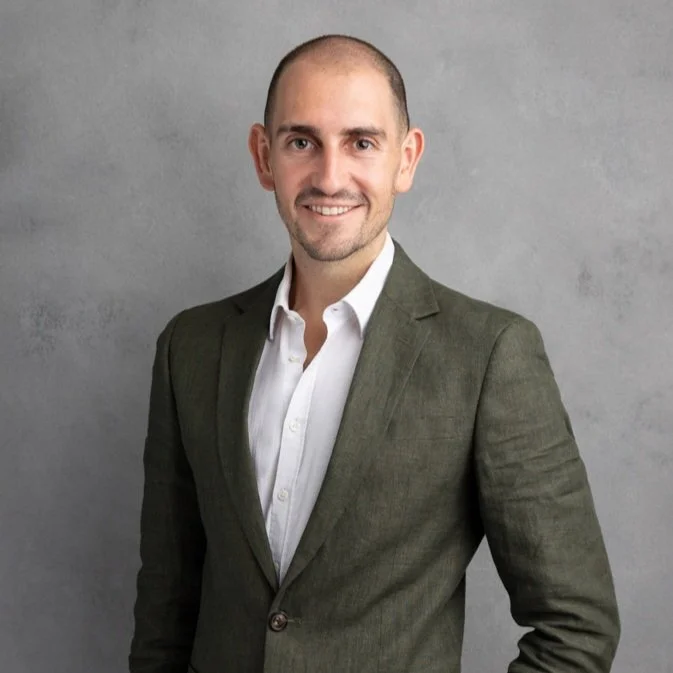 A smiling man with a shaved head and light beard, dressed in a white shirt and dark green blazer, standing against a gray background.