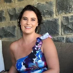 A woman with dark hair wearing a blue floral one-shoulder dress, smiling, sitting against a stone wall.