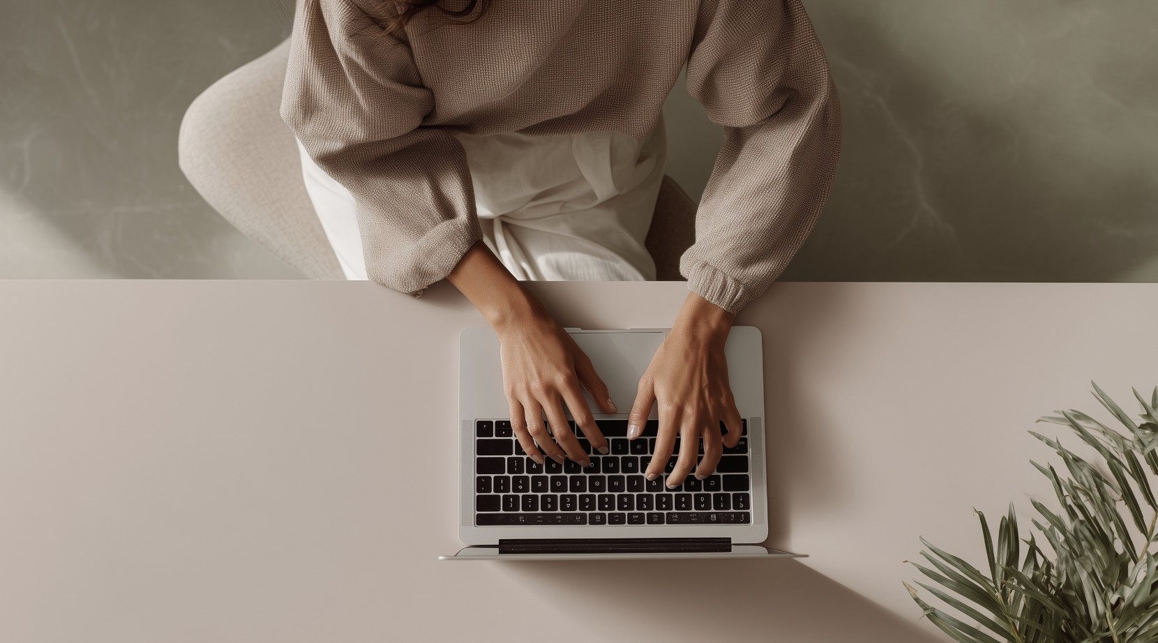 A person sitting at a desk using a laptop, viewed from above. The person is wearing a beige sweater and white pants, and there is a green plant on the right side of the desk.
