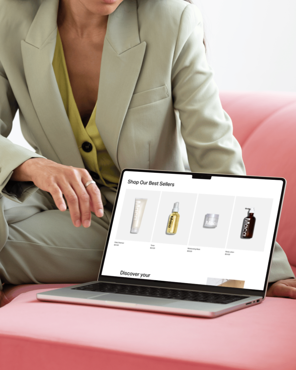 A person in a light-colored blazer with a yellow shirt underneath sitting on a pink couch and viewing a laptop screen displaying an online beauty product store with various skincare and makeup products.