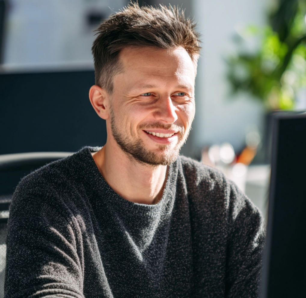 A young man with short brown hair and a beard smiling at a computer in a bright, modern office.