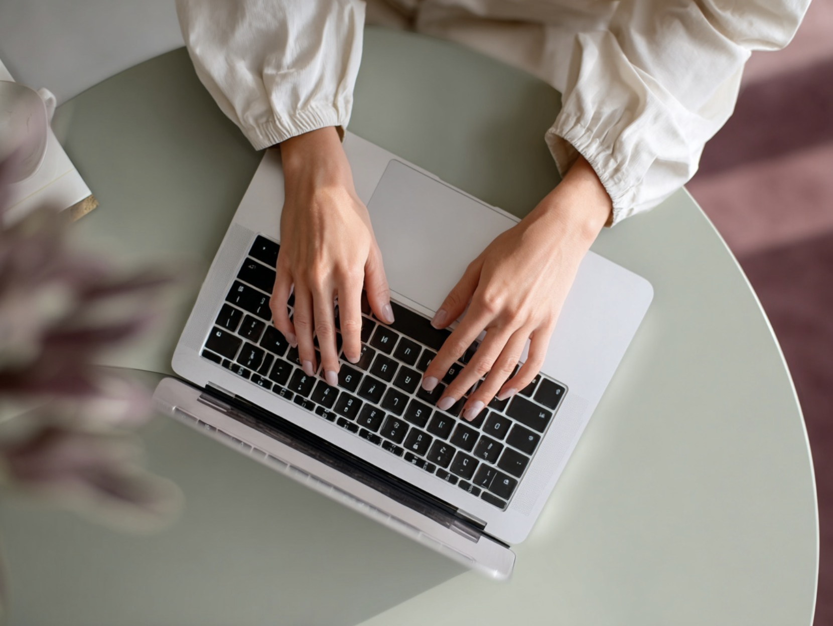 Person typing on a silver laptop with black keyboard, sitting at a light green table.