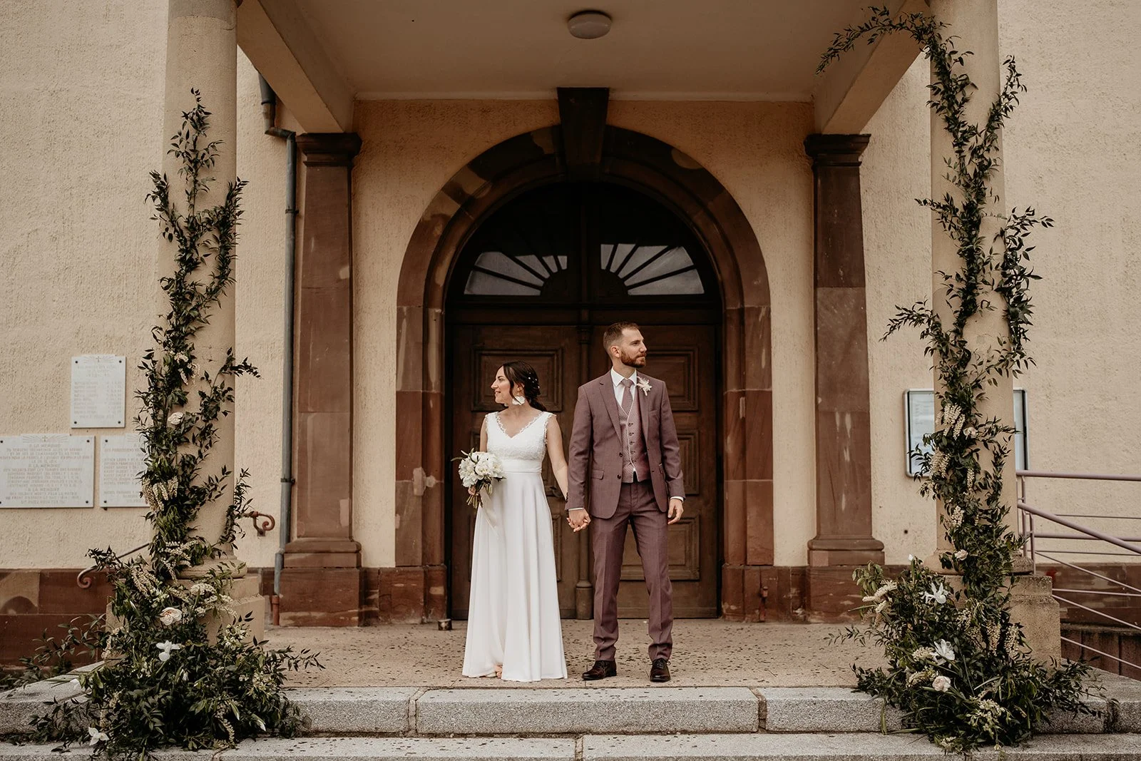 Un couple marié devant une grande porte en bois, décorée de feuillages et de fleurs, lors d'un mariage à l'extérieur. Design mariage France, Alsace.