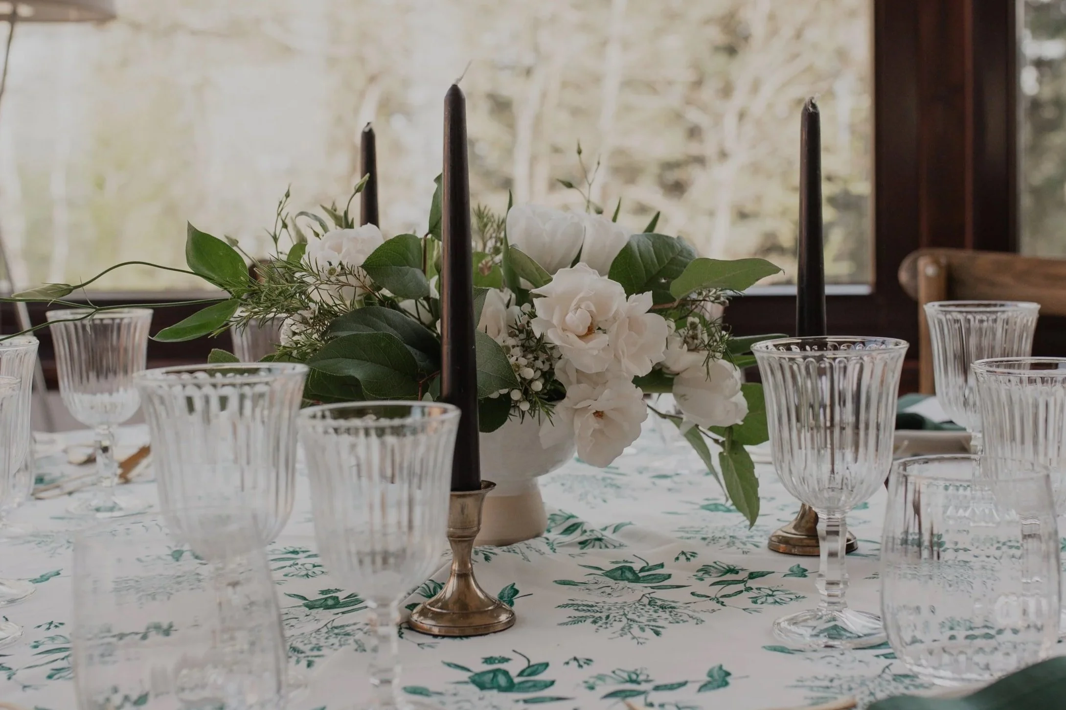 Table de dîner élégante avec une nappe à motif floral, une vase avec des fleurs blanches, des verres à vin en cristal et des chandeliers noirs pour un mariage style vintage British. Conseil mariage, coach mariage.