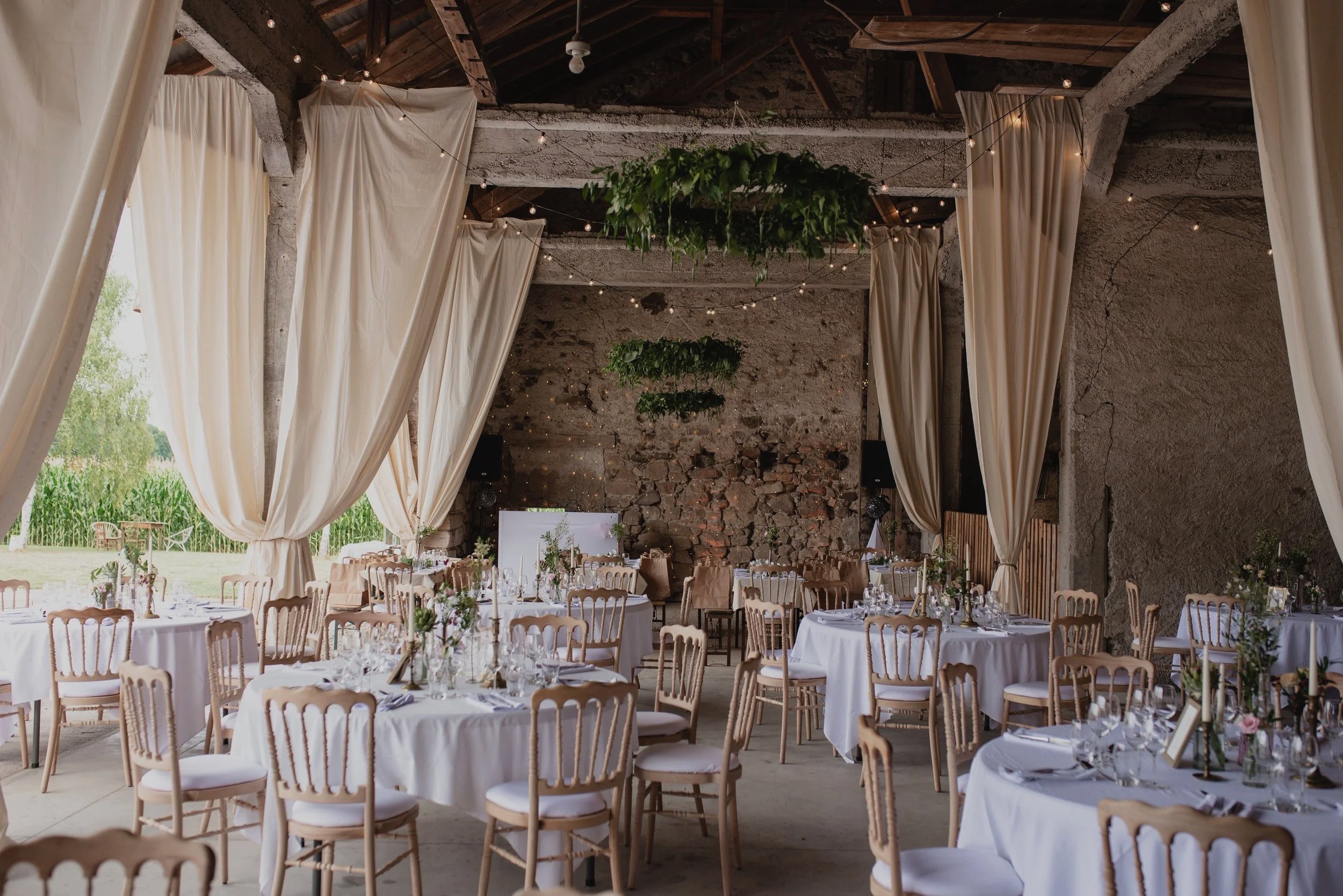 Salle de réception décorée pour un mariage, avec tables rondes, chaises en bois, décoration florale, guirlandes lumineuses dans une Ferme en Alsace avec murs en pierre et vue extérieure verdoyante.