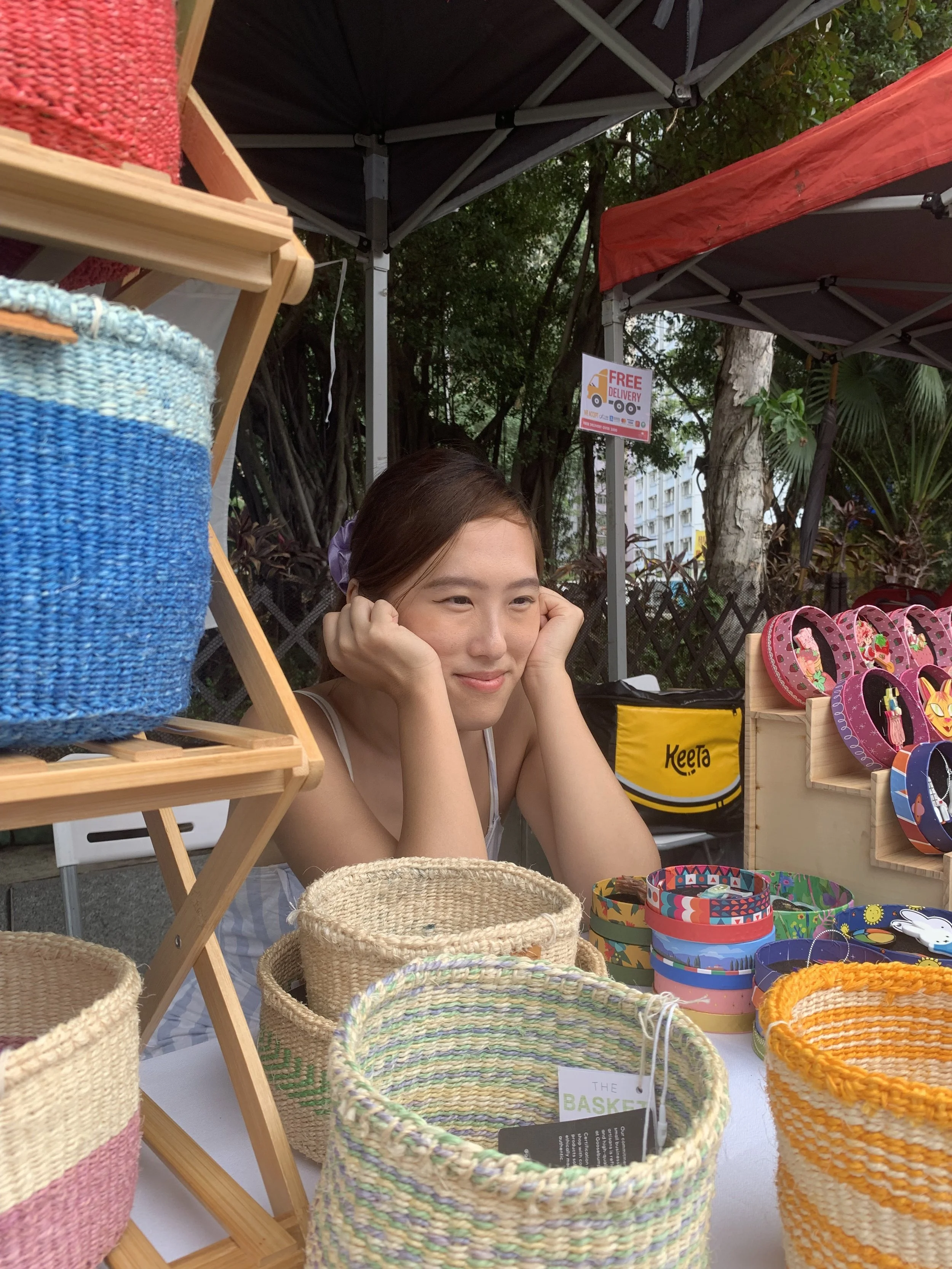 A young woman with brown hair, smiling and resting her head on her hands, sitting at a market stall selling woven baskets and colorful accessories. She is under a black canopy with trees and buildings in the background.