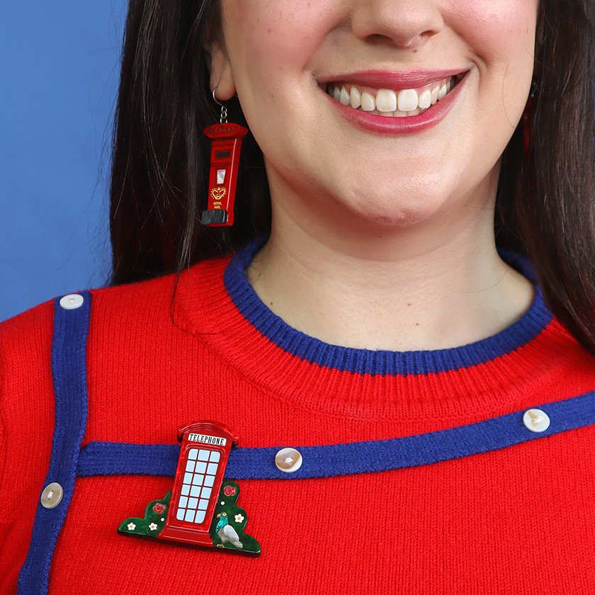 A brooch shaped like a vibrant red telephone booth, surrounded by green foliage with floral details, hand-assembled using resin, representing London’s iconic charm.