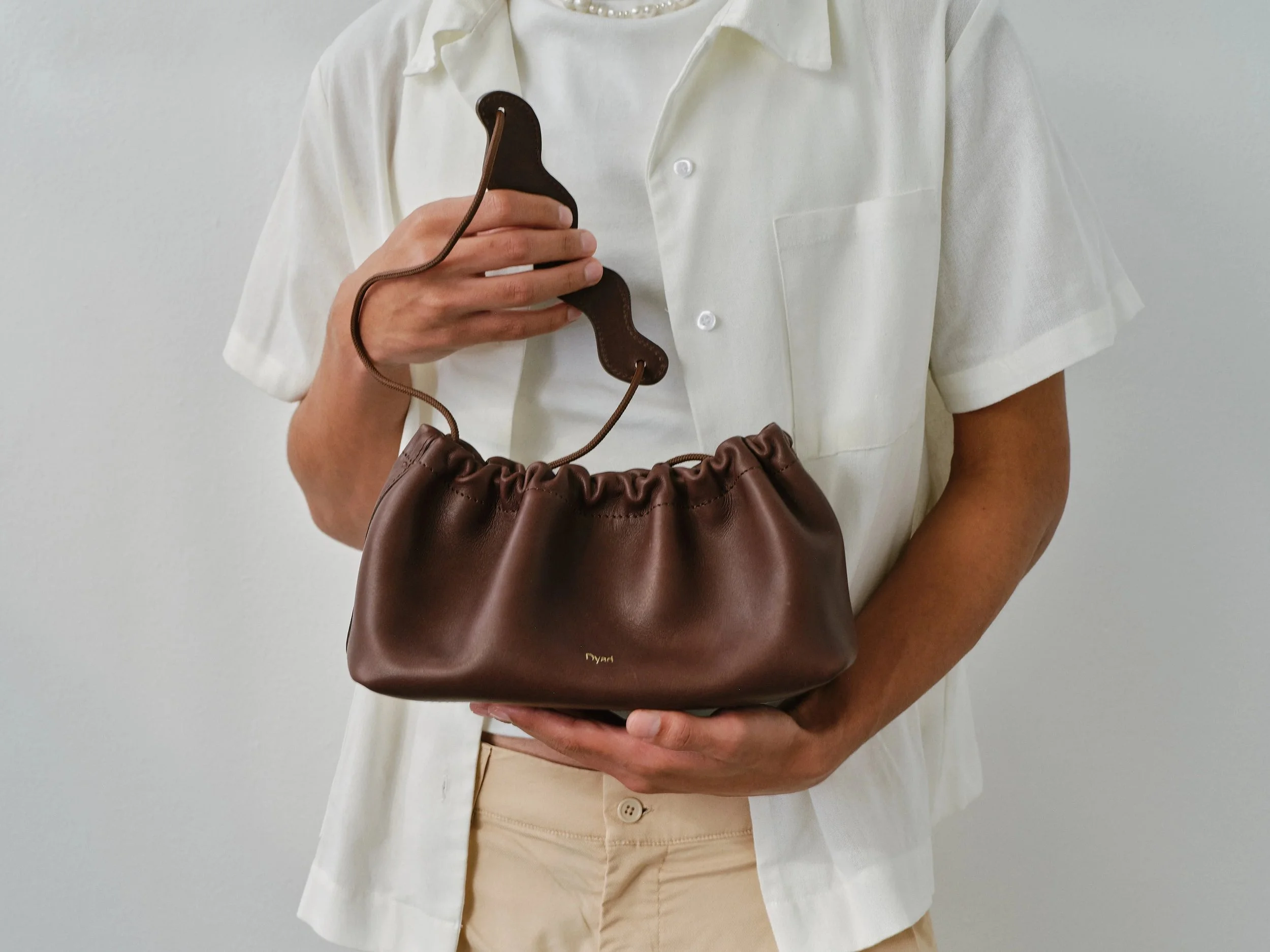 Chocolate brown pleated canvas shoulder bag by Project Dyad with adjustable strap and minimalist silhouette.