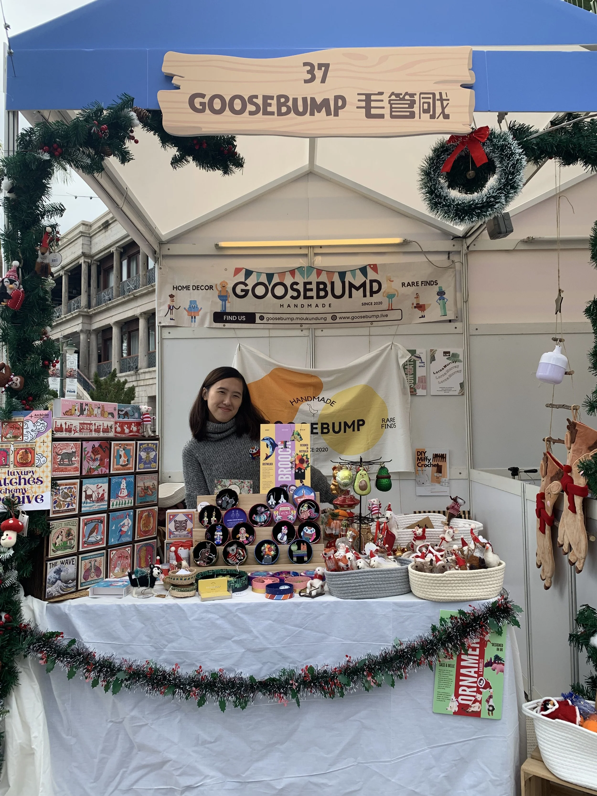 Woman selling handmade holiday decorations and accessories at a market stall decorated with Christmas garland and ornaments.