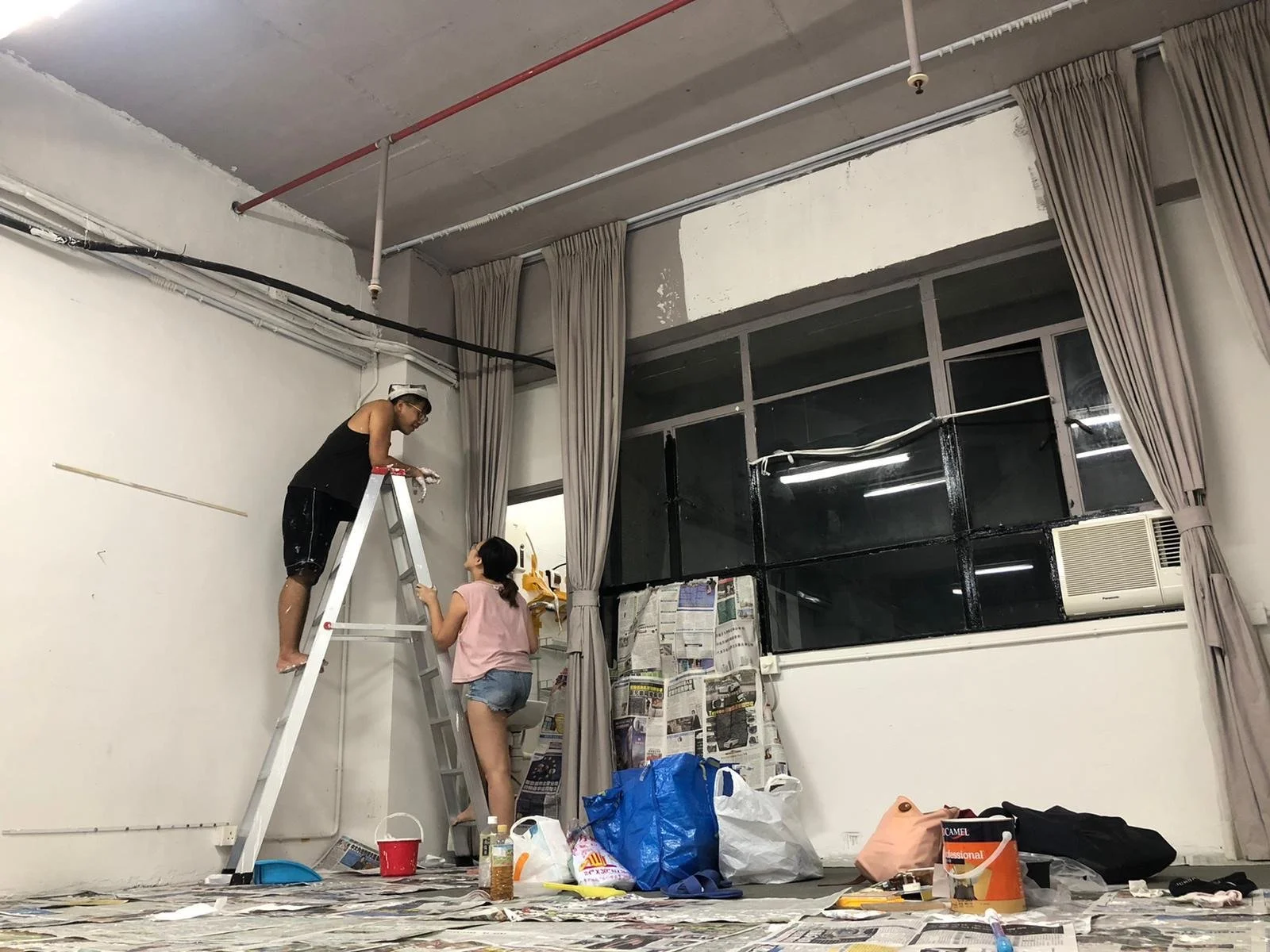 Two women painting a wall in a room; one standing on a ladder and the other on the floor assisting.
