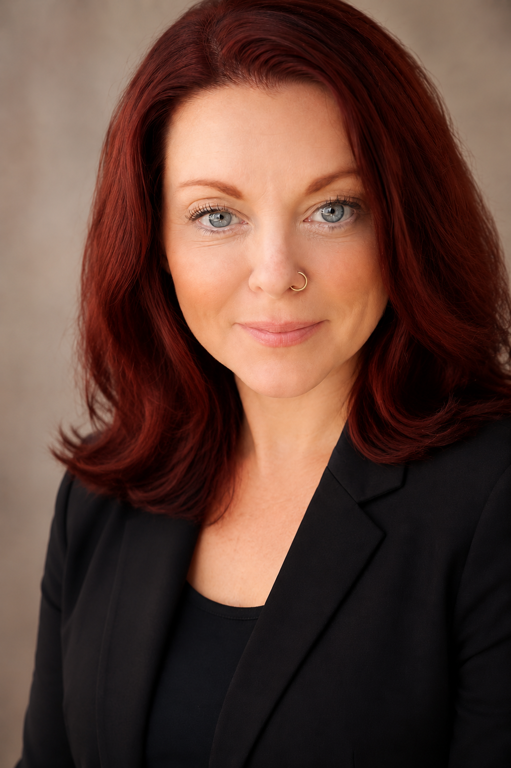 A woman with shoulder-length red hair, blue eyes, and a nose ring, wearing a black blazer and top, smiling softly at the camera. massage therapist Amy Marie DiFranco, reiki practitioner, holistic wellness practitioner, wome circle facilitator