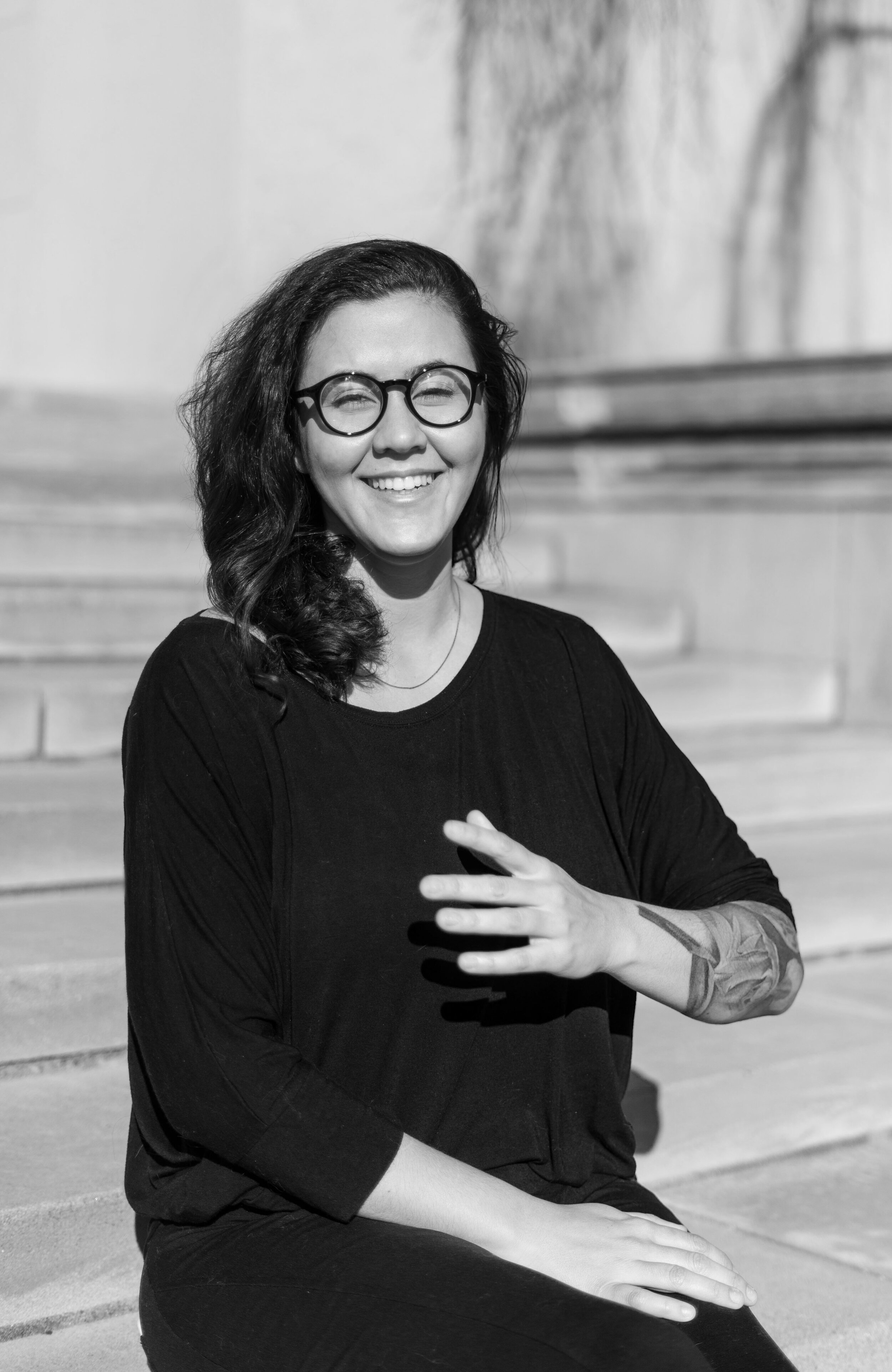 Person with long hair and glasses sitting on stone steps outdoors.