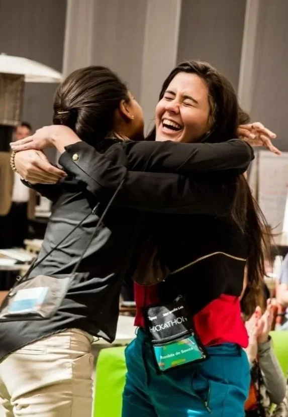 Two people wearing event badges hugging and smiling in a busy indoor setting.